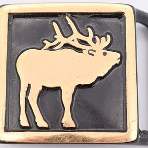 May include: A rectangular belt buckle featuring a gold-coloured elk design set against a black background. The buckle has a gold border and a black frame and loop. The elk is depicted standing with its head turned to the right.