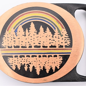 May include: A copper-colored belt buckle with a black handle. The circular face features a rainbow over a forest scene, with a mirrored reflection below. The buckle is a nature-themed accessory.