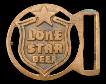 May include: A brass belt buckle featuring a central shield shape with a star above it. The text LONE STAR BEER is embossed in the center of the shield. The buckle has a circular frame with an integrated rectangular loop on the right side.