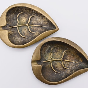 May include: Two brass leaf-shaped ashtrays with a raised leaf design. The ashtrays are a dark gold color and have a slightly textured surface.