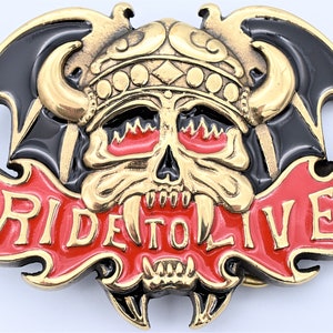 May include: Gold-tone belt buckle with a skull wearing a helmet and wings. The skull has red eyes with flames and a red banner with the words "Ride To Live".