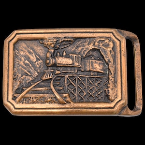 May include: A copper-colored rectangular belt buckle featuring a detailed relief of a steam train crossing a trestle bridge. The design includes mountains, a tunnel, and billowing smoke, with a classic Western theme.