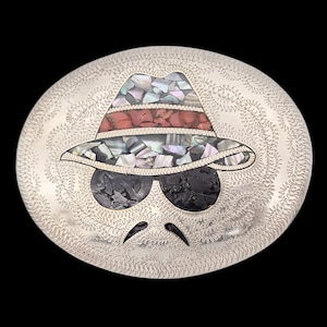 May include: Oval silver belt buckle with an inlaid design of a fedora hat, sunglasses, and a mustache. The hat features red, iridescent, and black stone inlays. The buckle has a detailed engraved border and is set against a black background.