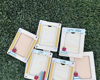 May include: Wooden picture frames with pencil and apple accents. Some frames have the names "Levi" and "Lian", and the phrase "Fifth Grade". The frames are arranged on a green grassy surface.