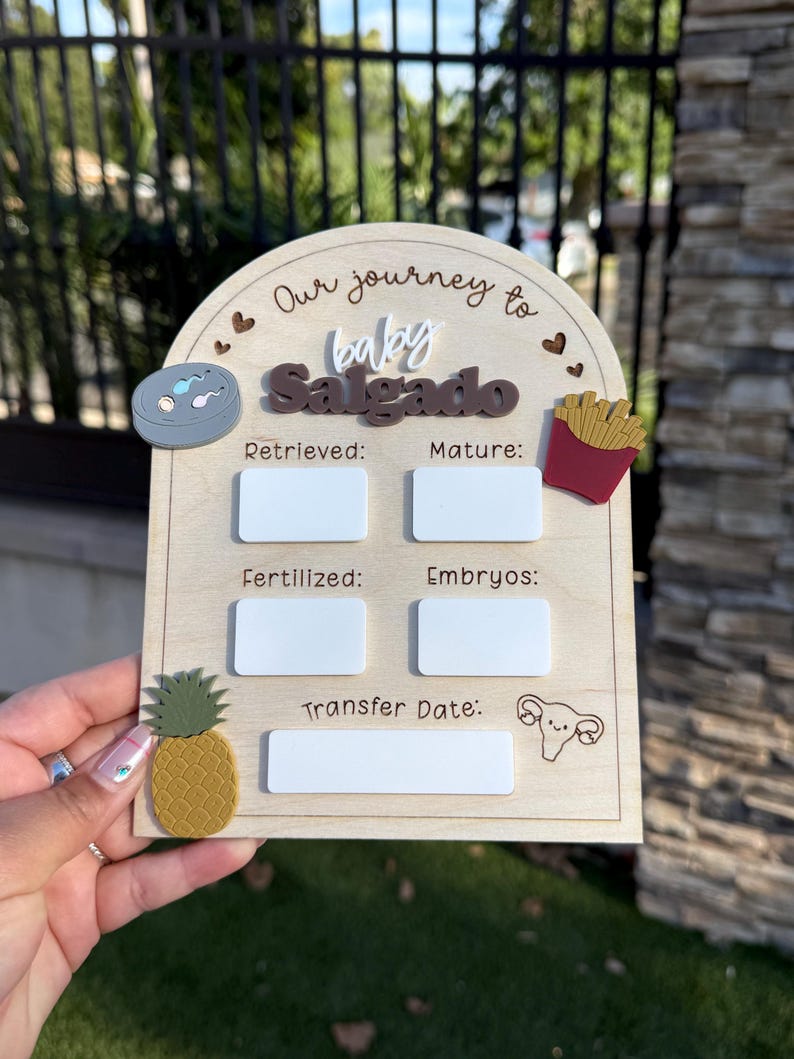 May include: An arched wooden IVF milestone tracker sign with the text Our journey to baby Salgado. It features white acrylic rectangles for writing data under the labels Retrieved, Mature, Fertilized, Embryos, and Transfer Date, with decorative icons.