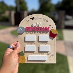 May include: An arched wooden IVF journey milestone sign with the text Our IVF journey to baby BINGHAM. It features five white rectangular dry-erase sections labeled Retrieved, Mature, Fertilized, Embryos, and Transfer Date, with decorative icons.