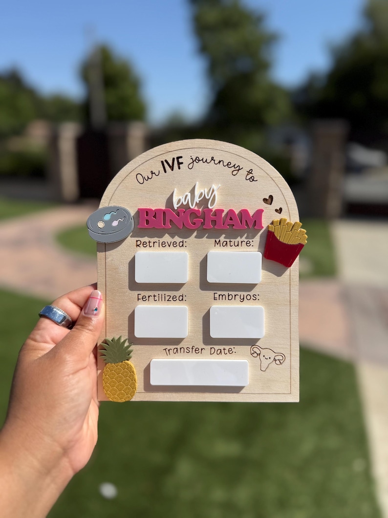 May include: An arched wooden IVF journey milestone sign with the text Our IVF journey to baby BINGHAM. It features five white rectangular dry-erase sections labeled Retrieved, Mature, Fertilized, Embryos, and Transfer Date, with decorative icons.