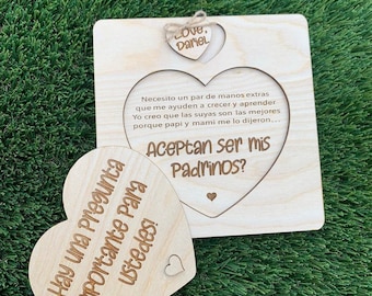 May include: Wooden heart-shaped sign with the text "Hay una pregunta importante para ustedes!" and a smaller wooden heart-shaped sign with the text "Aceptan ser mis Padrinos?"  The larger heart is attached to a square wooden sign with the text "Necesito un par de manos extras que me ayuden a crecer y aprender Yo creo que las suyas son las mejores porque papi y mami me lo dijeron... Love, Dariel"