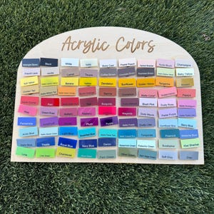 May include: A semi-circular wooden display board with the text Acrylic Colors printed at the top. It features a grid of numerous small rectangular acrylic swatches in a wide variety of colors, each labeled with its specific color name in black text.