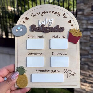 May include: An arched wooden sign titled Our journey to baby Salgado with white acrylic rectangles for writing data. It features icons of a petri dish, french fries, a pineapple, and a uterus, with labels for Retrieved, Mature, Fertilized, Embryos, and Transfer Date.
