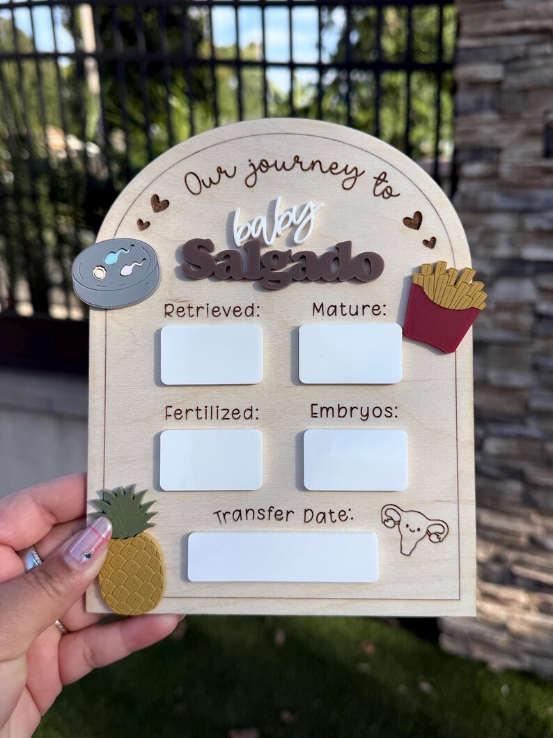 May include: An arched wooden sign titled Our journey to baby Salgado with white acrylic rectangles for writing data. It features icons of a petri dish, french fries, a pineapple, and a uterus, with labels for Retrieved, Mature, Fertilized, Embryos, and Transfer Date.