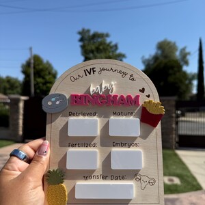 May include: An arched wooden sign with the text Our IVF journey to baby BINGHAM. It features white rectangular spaces for writing data next to labels for Retrieved, Mature, Fertilized, Embryos, and Transfer Date, with icons of a petri dish, fries, a pineapple, and a uterus.