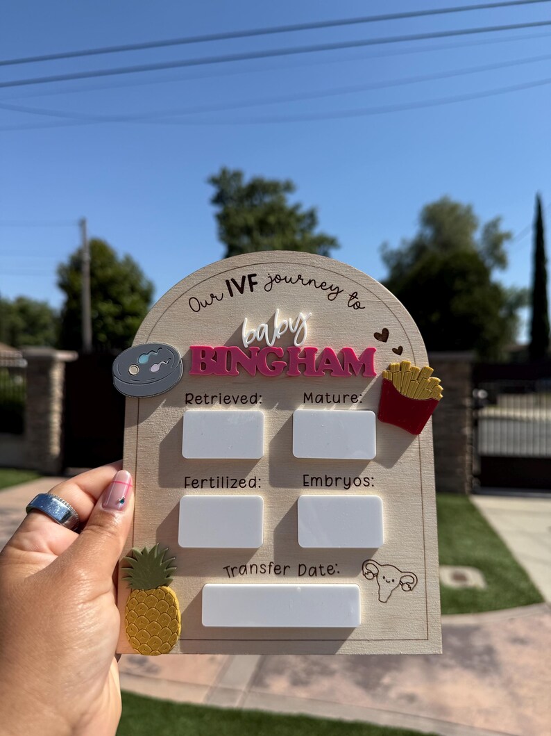 May include: An arched wooden sign with the text Our IVF journey to baby BINGHAM. It features white rectangular spaces for writing data next to labels for Retrieved, Mature, Fertilized, Embryos, and Transfer Date, with icons of a petri dish, fries, a pineapple, and a uterus.