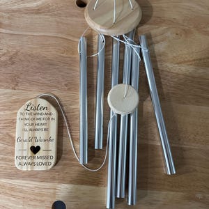 Pode incluir: Um móbile de vento de madeira com um tubo de carrilhão de metal prateado. A placa de madeira diz "Listen to the wind and think of me for in your heart I'll always be Gerald Warnke Forever missed Always loved".