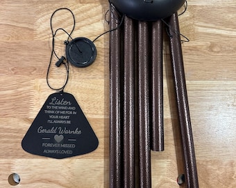 May include: A black wind chime with a triangular wooden plaque that reads "Listen to the wind and think of me for in your heart I'll always be Gerald Warnke Forever missed Always loved". The chime has six metal tubes that are a dark brown color.