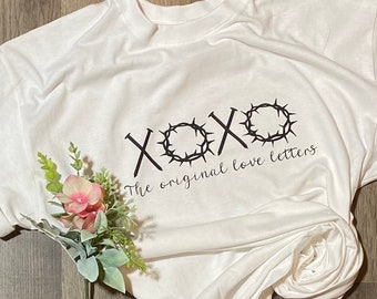 May include: White t-shirt with a black graphic design of the word "XOXO" with a crown of thorns around the "O". The text "The original love letters" is below the graphic.