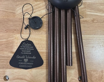 May include: A black wind chime with bronze-colored tubes. The chime has a circular top and a black tag with the text "Listen to the wind and think of me for in your heart I'll always be Gerald Warnke Forever missed Always loved."