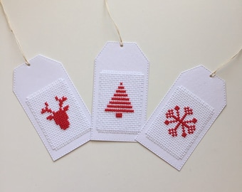 May include: Three white gift tags with cross-stitched holiday designs: a red reindeer, a Christmas tree, and a snowflake. Each tag has a stitched white pocket and a natural twine string for hanging.