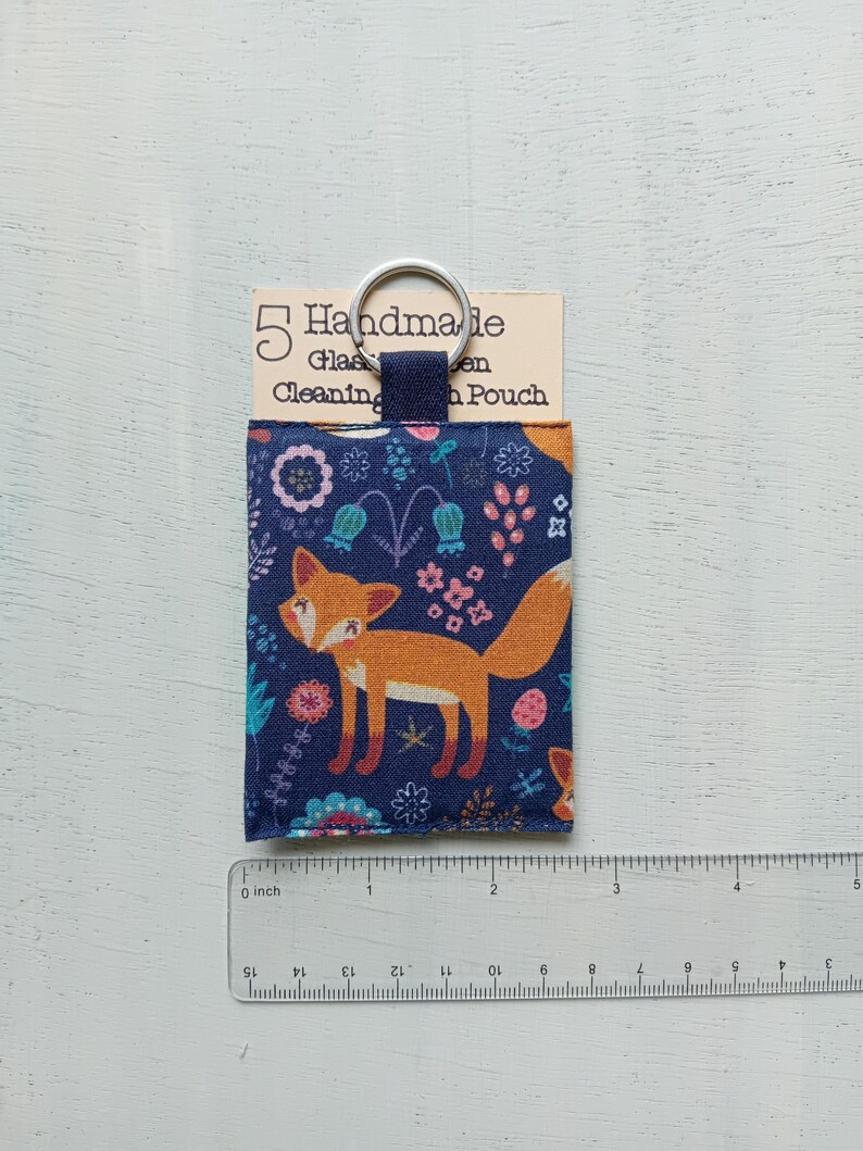 May include: A rectangular fabric pouch featuring a navy blue background with a repeating orange fox and floral pattern. The pouch has a dark blue fabric loop attached to a silver metal keyring. A tag reads 5 Handmade Glass Cleaning Pouch.