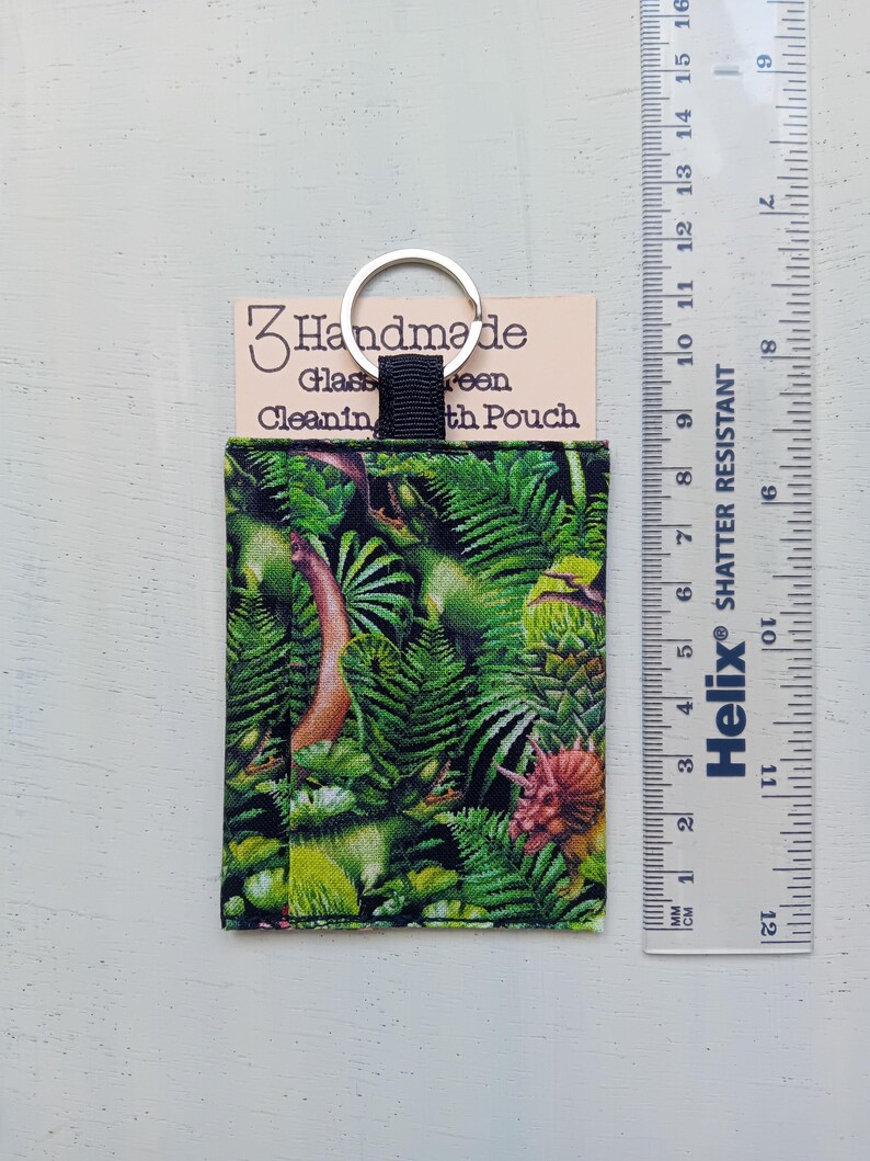 May include: A rectangular fabric pouch featuring a green dinosaur and fern print, attached to a silver metal keyring. A beige card behind the pouch reads "Handmade Glass Cleaning Cloth Pouch". A ruler shows the pouch is approximately 3.5 inches tall.