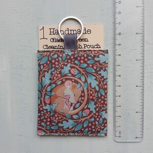 May include: A rectangular fabric pouch with a brown background featuring a pattern of teal oak leaves and pink berries, with a central circular graphic of a sleeping mouse. A silver keyring is attached to a gray tab. A ruler shows a height of 6 inches.
