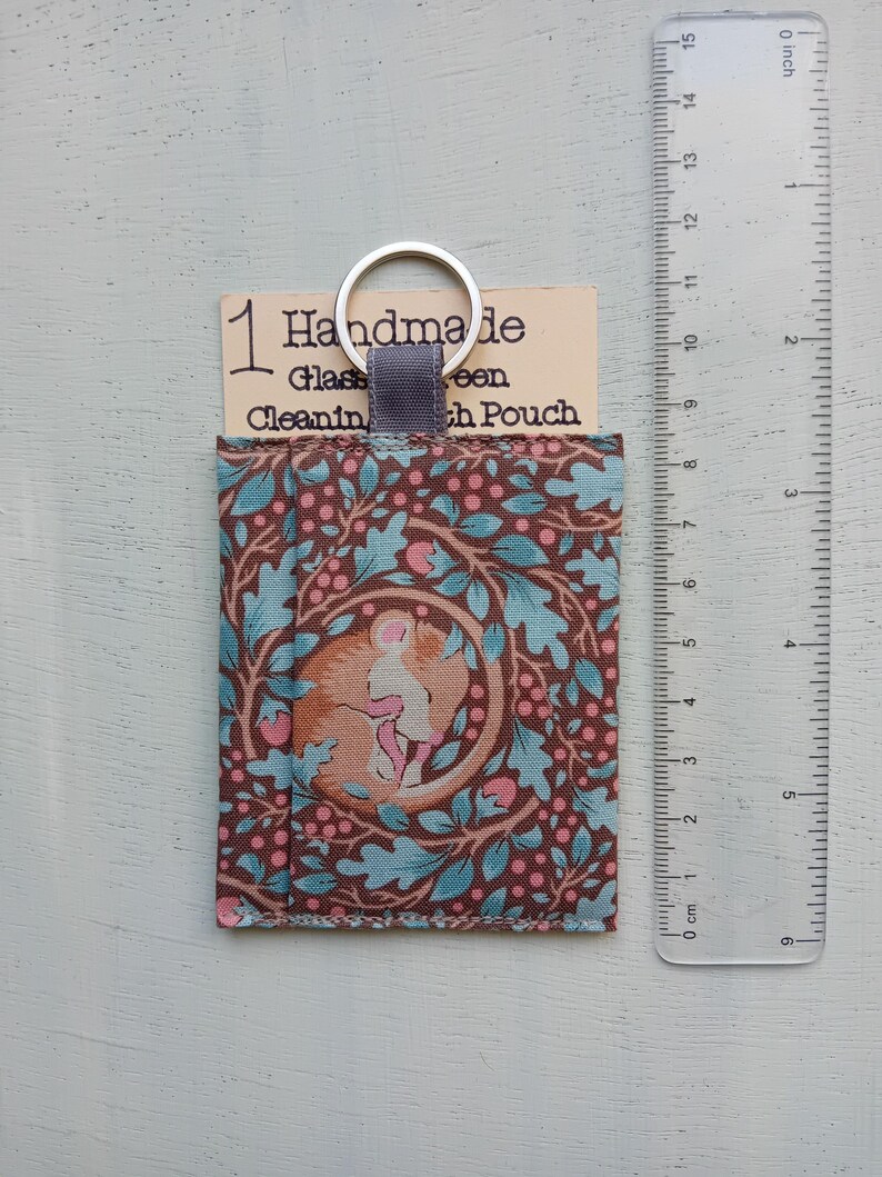 May include: A rectangular fabric pouch with a brown background featuring a pattern of teal oak leaves and pink berries, with a central circular graphic of a sleeping mouse. A silver keyring is attached to a gray tab. A ruler shows a height of 6 inches.