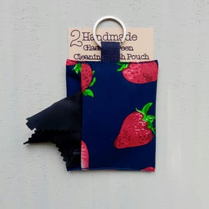 May include: A dark blue fabric pouch featuring a red strawberry pattern with green leaves. A black cleaning cloth protrudes from the side. The pouch has a metal keyring attached to a blue tab and a tag reading 2 Handmade Glass Cleaning Cloth Pouch.