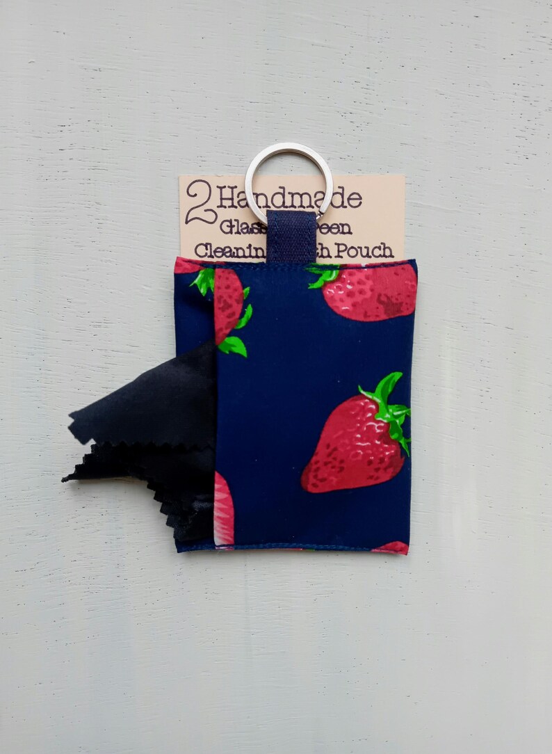 May include: A dark blue fabric pouch featuring a red strawberry pattern with green leaves. A black cleaning cloth protrudes from the side. The pouch has a metal keyring attached to a blue tab and a tag reading 2 Handmade Glass Cleaning Cloth Pouch.