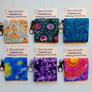May include: Six square fabric pouches labeled 1 through 6, each titled Handmade Glasses/screen Cleaning Cloth Pouch. The pouches feature various patterns: orange flowers, dark floral, blue marble, starry night, purple flowers, and a pink tie-dye design.