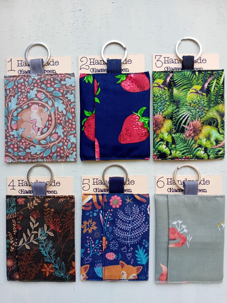 May include: Six rectangular fabric pouches with metal key rings, each attached to a card labeled 1 through 6. The pouches feature various prints including a sleeping animal, strawberries, dinosaurs, floral patterns, a fox, and a small animal on sage green.