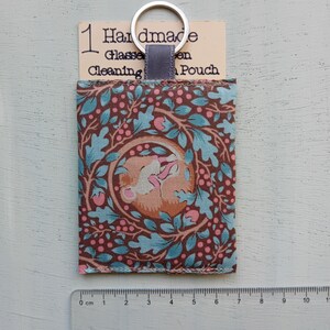 May include: A rectangular fabric glasses cleaning pouch featuring a brown, light blue, and pink botanical pattern with a sleeping animal. A silver metal keyring is attached to the top. A ruler shows the pouch is approximately 4.5 inches tall.