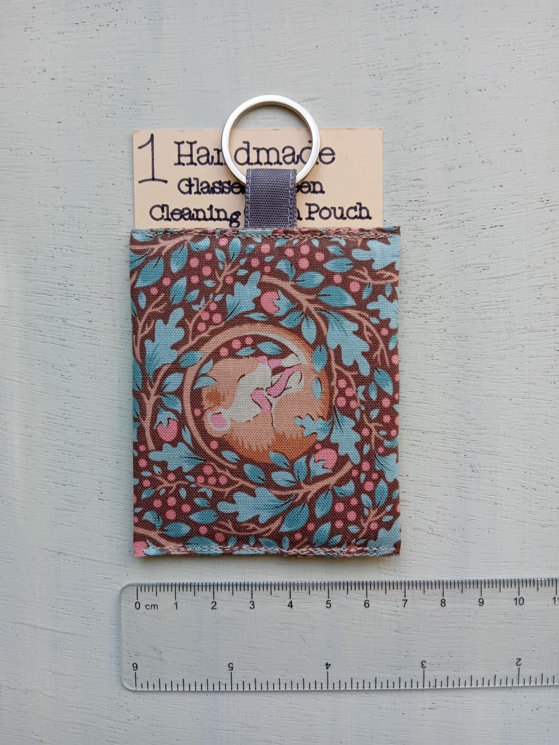 May include: A rectangular fabric glasses cleaning pouch featuring a brown, light blue, and pink botanical pattern with a sleeping animal. A silver metal keyring is attached to the top. A ruler shows the pouch is approximately 4.5 inches tall.