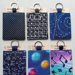 May include: Six rectangular fabric keychains with metal rings, each attached to a card labelled Handmade Glasses Seen. Designs include black with white dots, a periodic table, blue with yellow stars, math equations, planets, and musical notes.