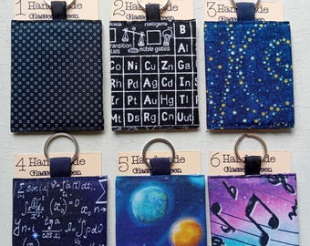 May include: Six rectangular fabric keychains with metal rings, each attached to a card labeled Handmade Glasses Seen. Designs include black with white dots, a periodic table, blue with yellow stars, math equations, planets, and musical notes.