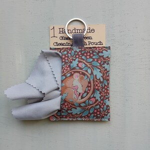 May include: A rectangular fabric pouch featuring a brown and teal pattern of a curled-up mouse surrounded by oak leaves and berries. A light gray microfiber cloth protrudes from the top. A silver metal keyring is attached to a gray fabric loop.