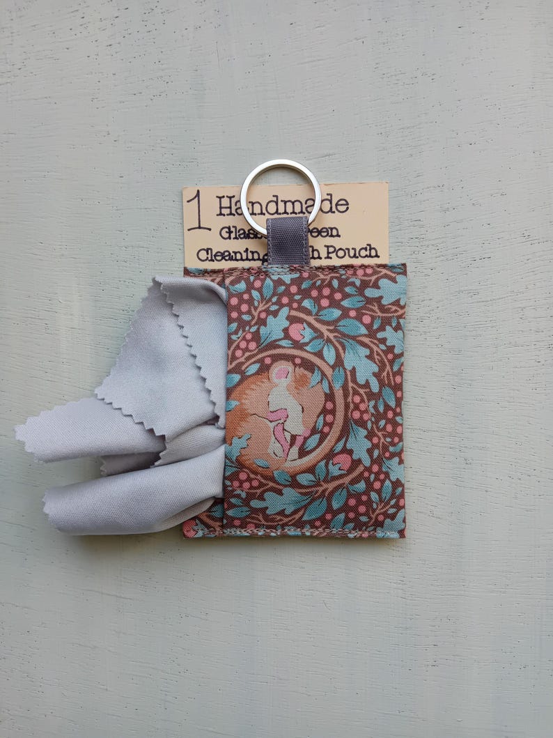 May include: A rectangular fabric pouch featuring a brown and teal pattern of a curled-up mouse surrounded by oak leaves and berries. A light gray microfiber cloth protrudes from the top. A silver metal keyring is attached to a gray fabric loop.