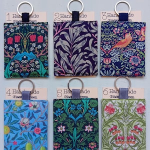 May include: Six rectangular fabric keychains with metal rings, each featuring distinct floral and botanical patterns in various colours including blue, green, purple, and red. Each keychain is attached to a card labelled with a number from one to six.