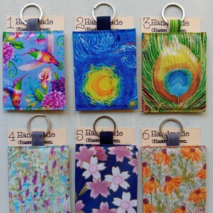 May include: Six handmade glasses cases with key rings. Each case features a unique fabric design: hummingbirds, Van Gogh's Starry Night, a peacock feather, lavender, pink flowers, and orange flowers. Each case has a tag with the words "Handmade" and "Glass".