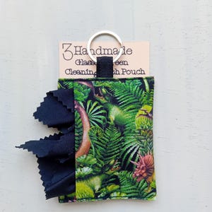 May include: A fabric pouch featuring a green dinosaur and jungle leaf print with a black cloth extending from the side. A silver metal keyring is attached to the top. The pouch is displayed on a card labeled 3 Handmade Glass Cleaning Cloth Pouch.