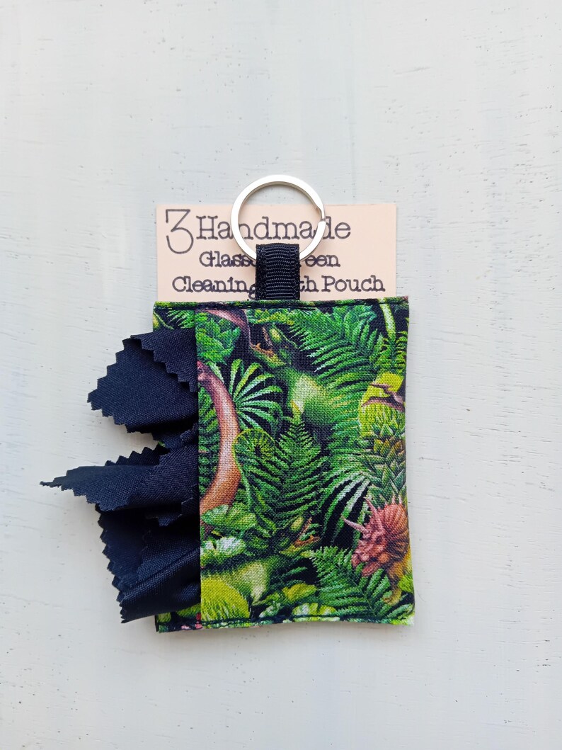May include: A fabric pouch featuring a green dinosaur and jungle leaf print with a black cloth extending from the side. A silver metal keyring is attached to the top. The pouch is displayed on a card labeled 3 Handmade Glass Cleaning Cloth Pouch.