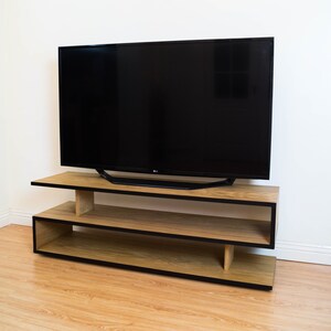 May include: A wooden TV stand with black trim and two shelves. The stand is made of light wood and has a modern design. A flat screen TV is sitting on top of the stand.