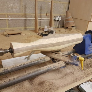 May include: A wooden object is being turned on a wood lathe. The object is made of two different types of wood, one light and one dark. The lathe is blue and has a yellow lever. The object is being turned into a smooth, rounded shape.