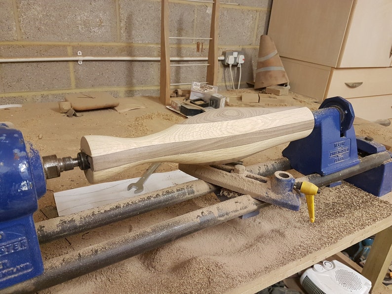 May include: A wooden object is being turned on a wood lathe. The object is made of two different types of wood, one light and one dark. The lathe is blue and has a yellow lever. The object is being turned into a smooth, rounded shape.