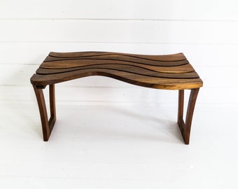 May include: A wooden bench with a wavy top and a dark brown finish. The bench has a simple design with two legs on each side.