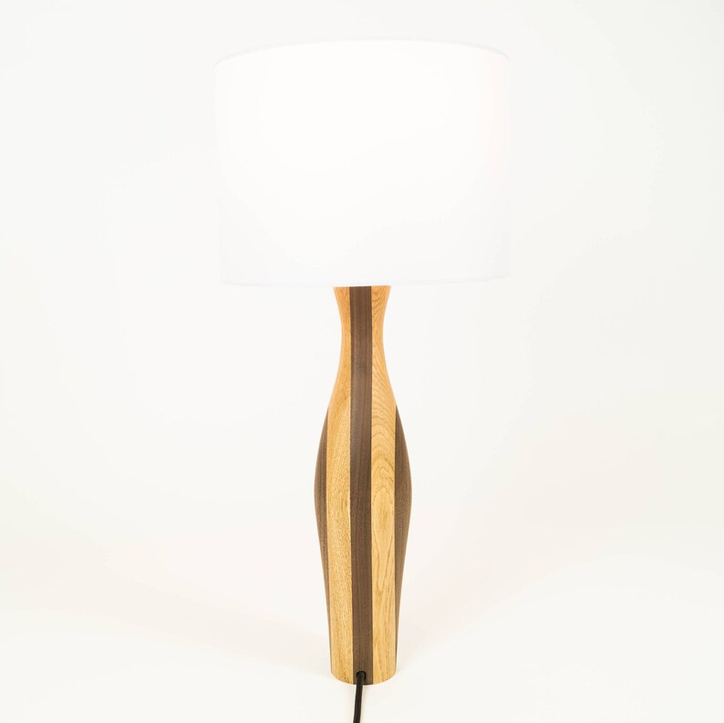 May include: A wooden table lamp with a white drum shade. The lamp base is made of alternating strips of light and dark wood.