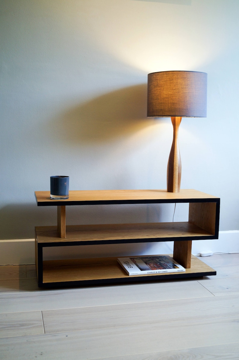 May include: A wooden shelf unit with black trim, featuring two shelves and a lamp with a gray fabric shade. A book titled "Eames" by Charles & Ray Eames is on the bottom shelf.