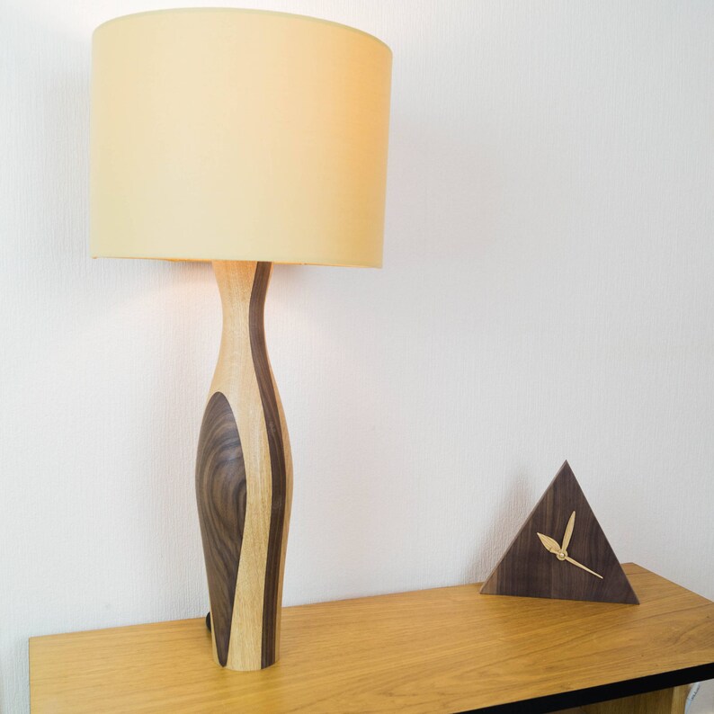 May include: A wooden table lamp with a beige lampshade and a wooden base with a unique swirl pattern. A small wooden triangle clock with gold hands is on the table.