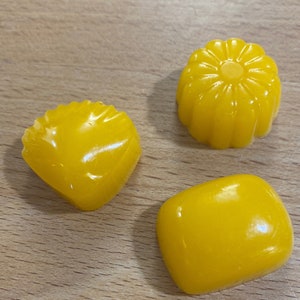 May include: Three yellow, handmade soap bars in different shapes. The soap bars are on a wooden surface.