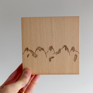 May include: A square, light-coloured wooden block featuring a mountain range etched along the bottom. The mountains are depicted with simple, angular lines. The block is held in a hand.