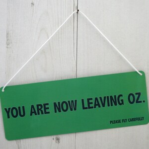 May include: A green sign with white string hanging from the top. The sign reads "You are now leaving Oz. Please fly carefully."